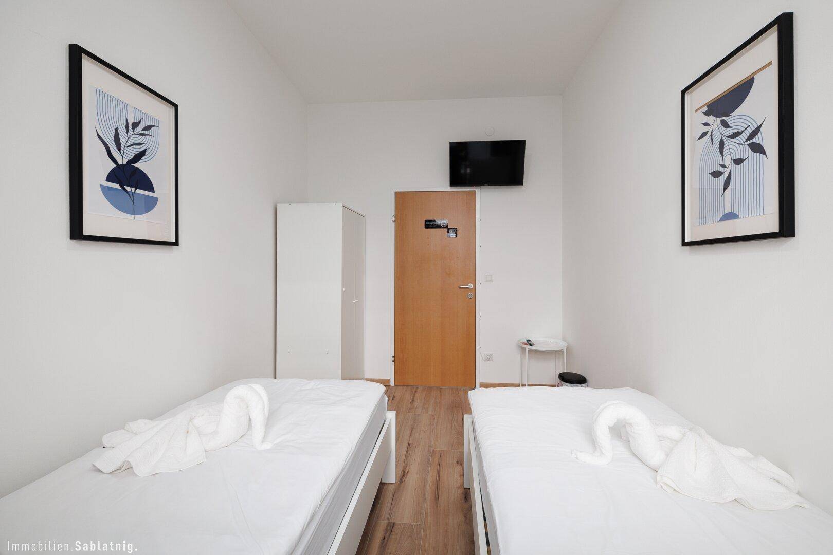Helles Schlafzimmer mit zwei Einzelbetten, Holzboden und einem an der Wand montierten Fernseher.