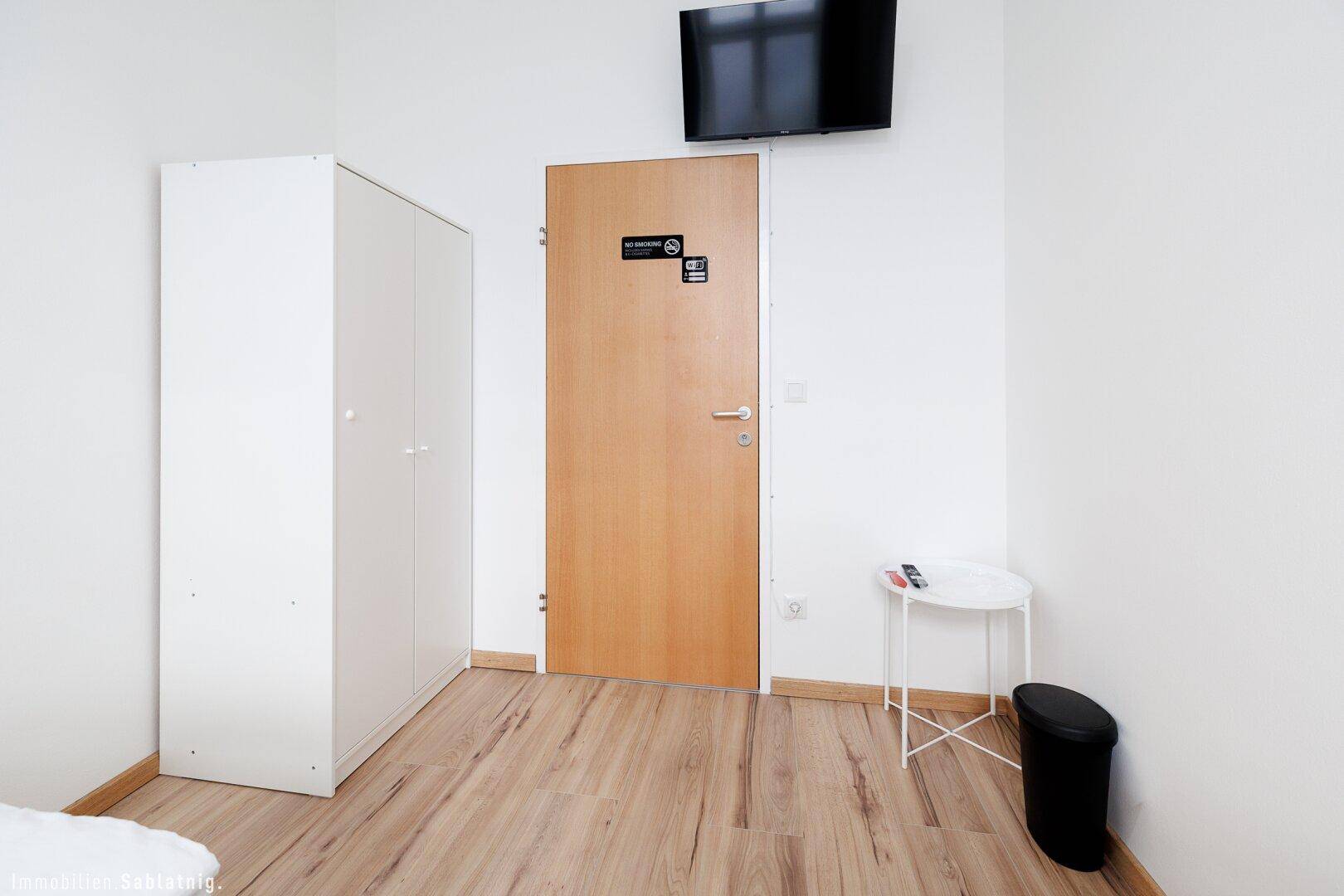 Bereich neben den Betten mit einem weißen Kleiderschrank, einer Holztür und einem an der Wand montierten Fernseher.