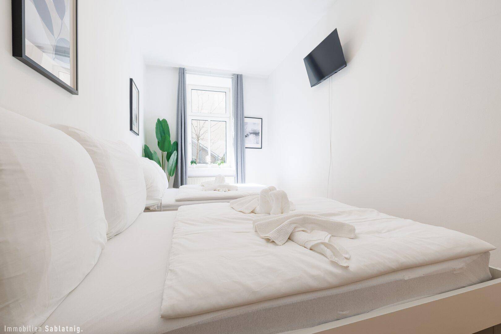 Helles Schlafzimmer mit zwei Einzelbetten, Holzboden und einem Fenster mit grauen Vorhängen.