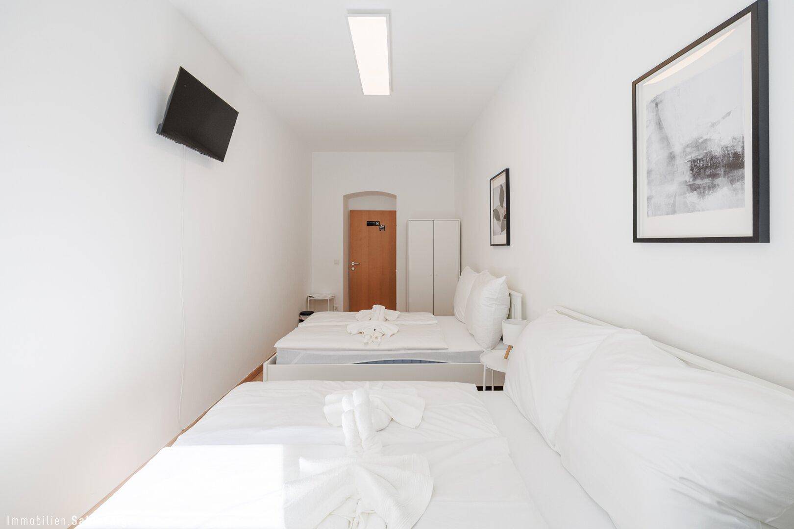 Helles Schlafzimmer mit zwei Einzelbetten, einem Wandfernseher und moderner Dekoration in schlichtem Design.