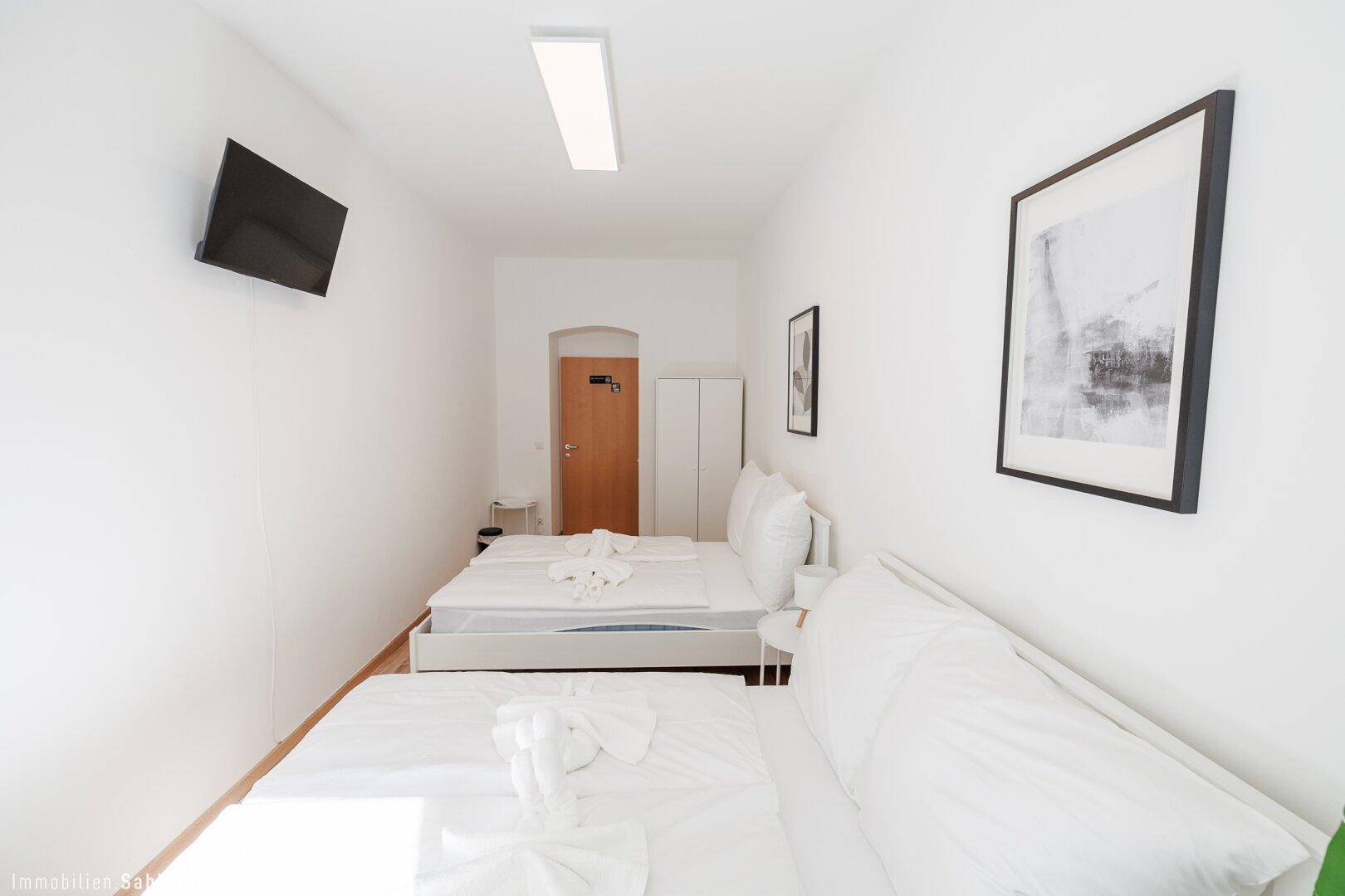 Schlichtes Schlafzimmer mit zwei Einzelbetten, einem Wandfernseher und minimalistischer Dekoration in Weißtönen.