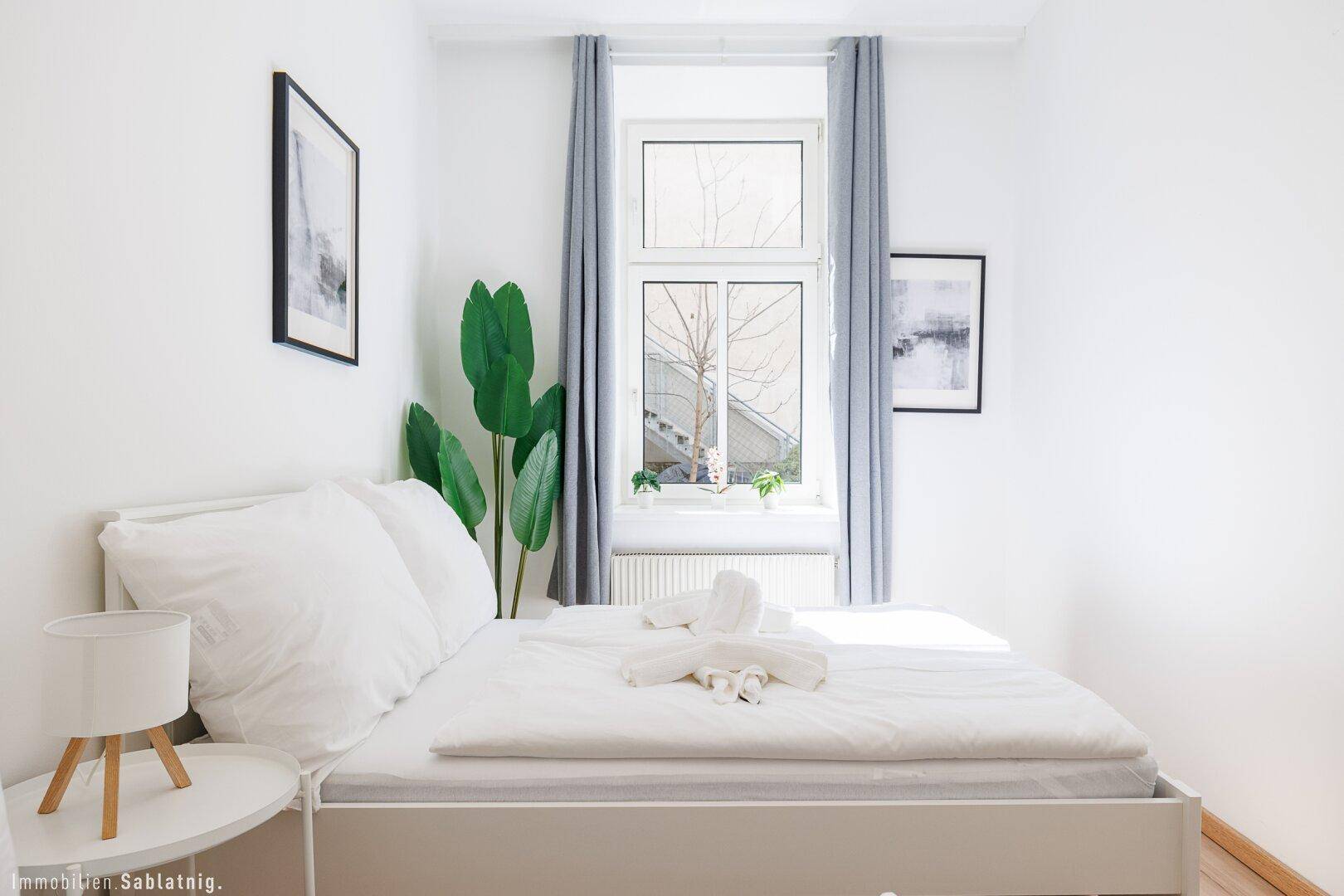 Gemütliches Schlafzimmer mit Doppelbett, großem Fenster und grüner Pflanzendekoration, hell und einladend.