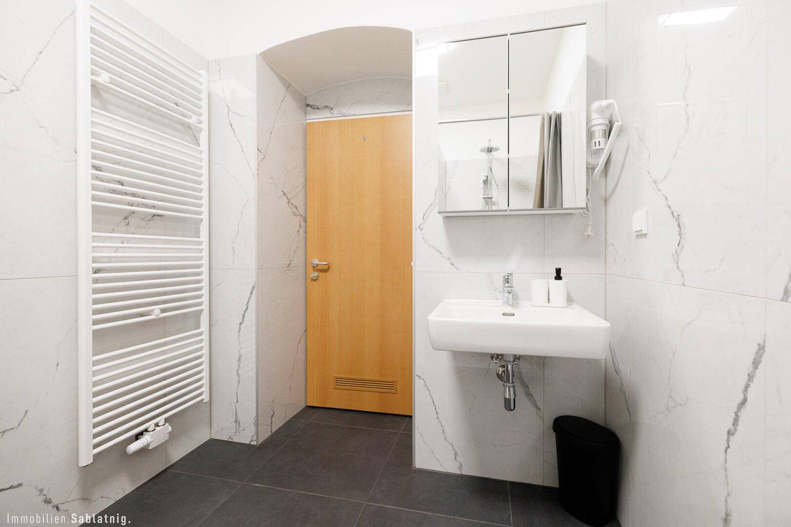 Modernes Badezimmer mit Handtuchheizkörper, Waschbecken und Spiegelschrank, alles in Weiß und Marmoroptik.