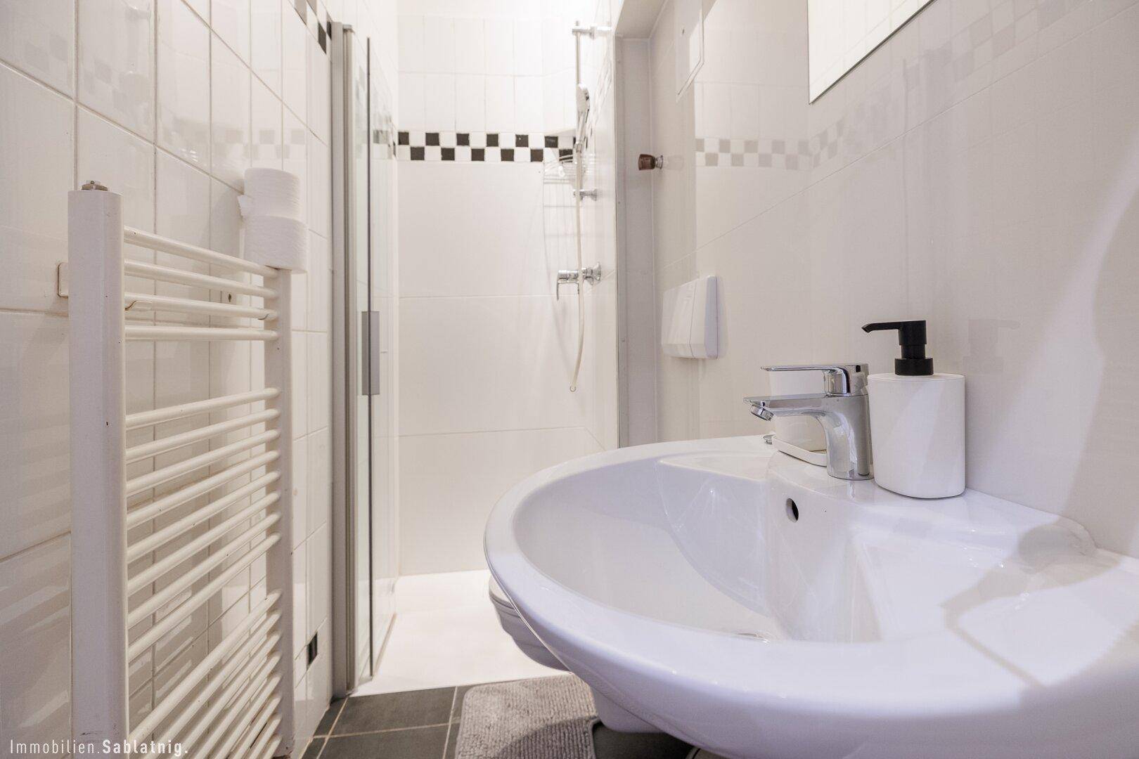 Nahaufnahme eines Waschbeckens in einem schmalen Badezimmer mit Duschbereich und Handtuchheizkörper.