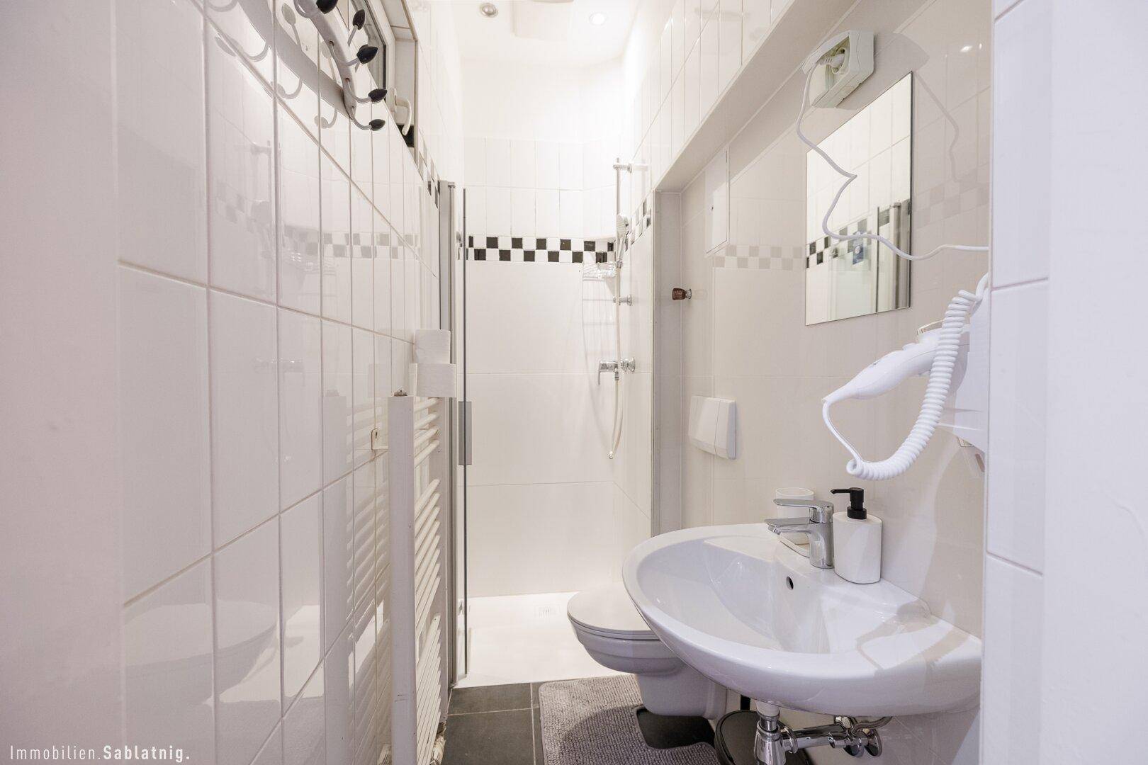 Schmaler, moderner Badezimmerbereich mit Dusche, Waschbecken und Wand-WC, alles in Weiß gehalten.