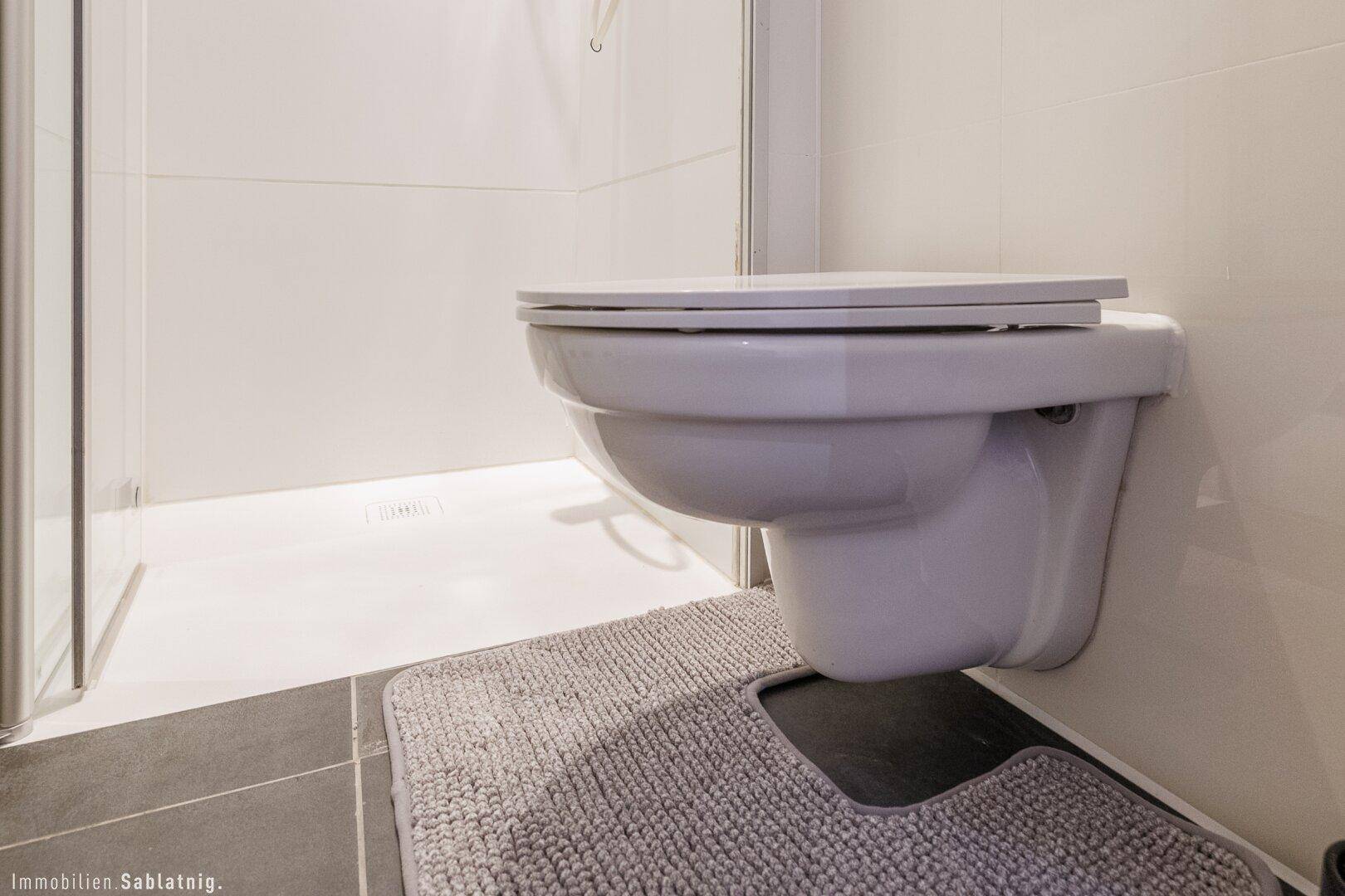 Wand-WC in einem modernen Badezimmer mit dunklem Fliesenboden und Duschbereich, sehr funktional.