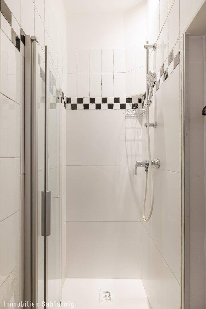 Duschbereich in einem schmalen Badezimmer mit weißen Fliesen und schwarzem Schachbrettmuster, modern gestaltet.