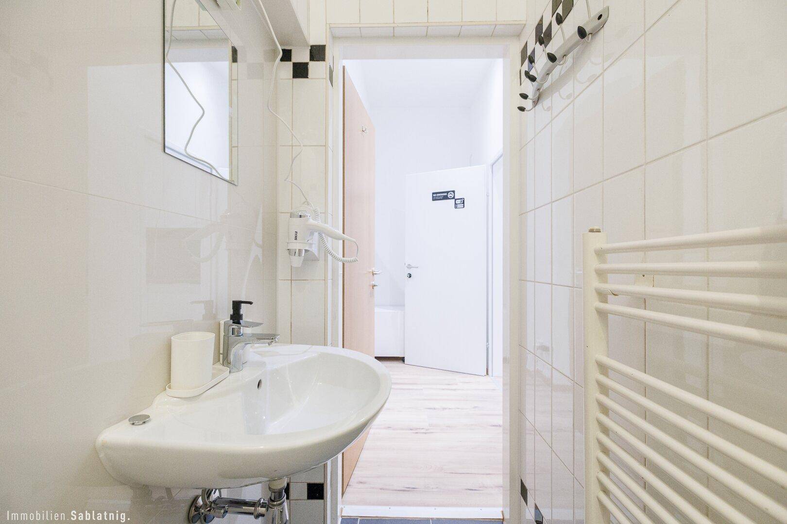 Modernes Badezimmer mit Waschbecken, Handtuchheizkörper und Blick in den Flur, hell und funktional.