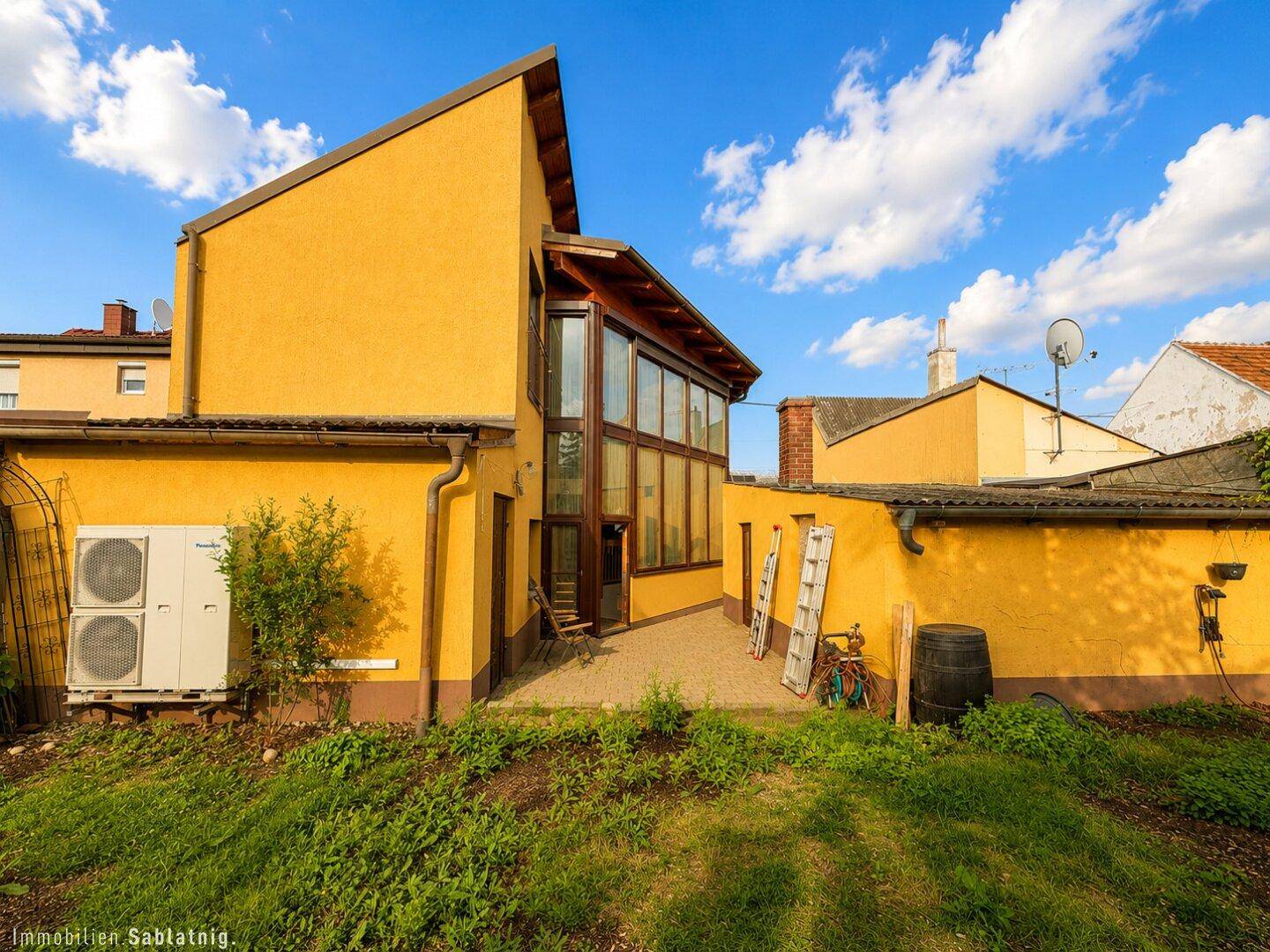 Rückseite des Hauses mit gelber Fassade, Klimaanlage und gepflastertem Bereich.