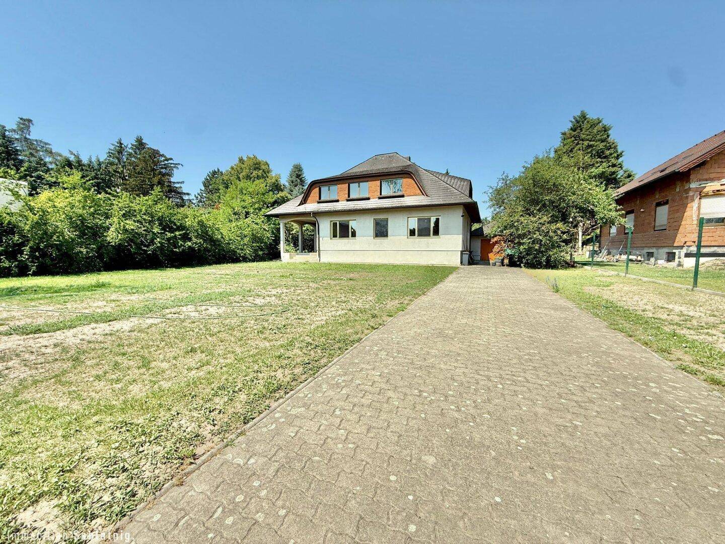Vorderansicht des Hauses mit gepflasterter Zufahrt, Rasenfläche und umgebenden Bäumen.
