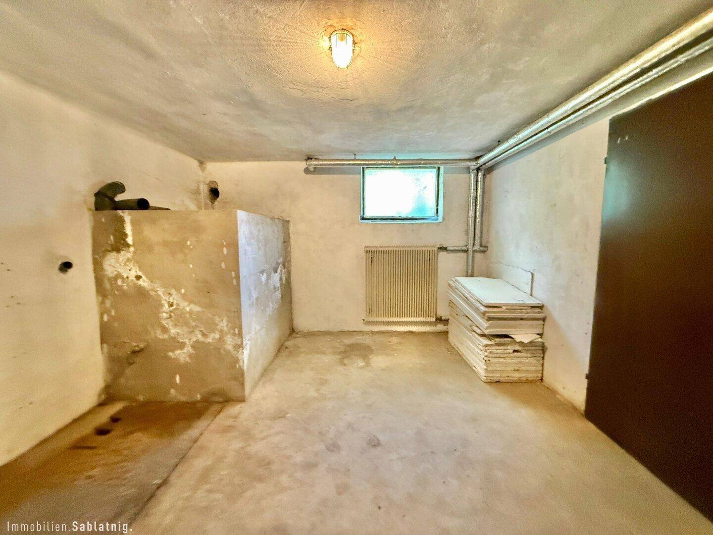 Unfertiger Kellerraum mit Betonboden, sichtbaren Rohren und kleinem Fenster.