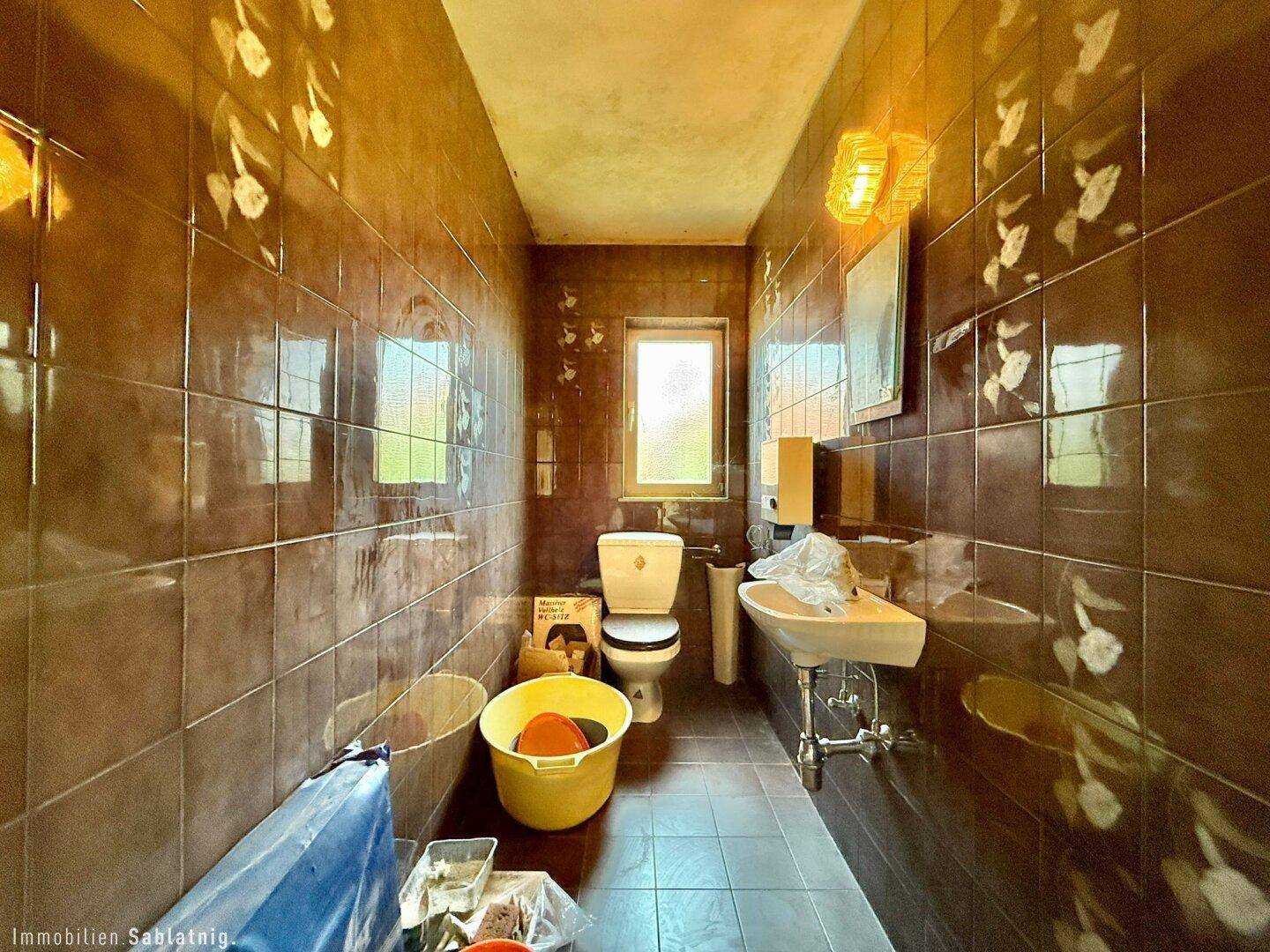 Separate Toilette mit braunen Fliesen, kleinem Waschbecken und Fenster.
