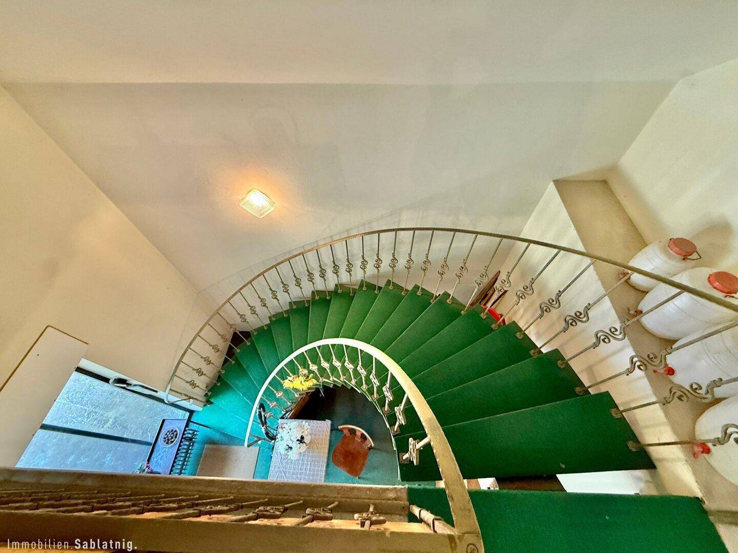 Wendeltreppe mit grünem Teppich und schmiedeeisernem Geländer.