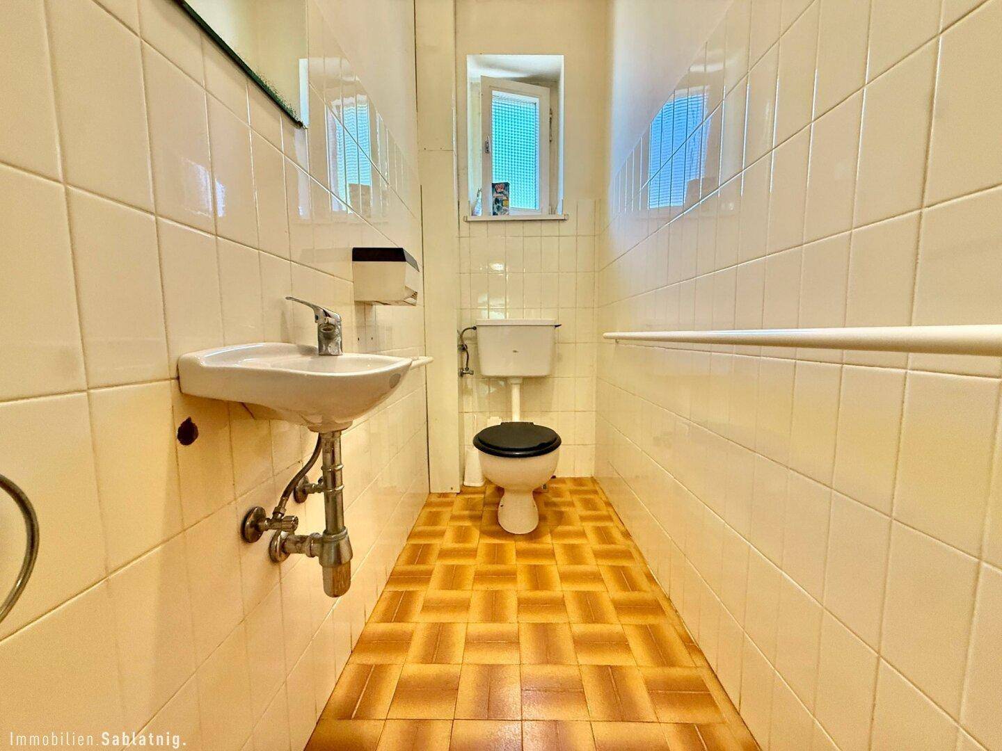 Separate Toilette mit weißen Fliesen, kleinem Waschbecken und Fenster.
