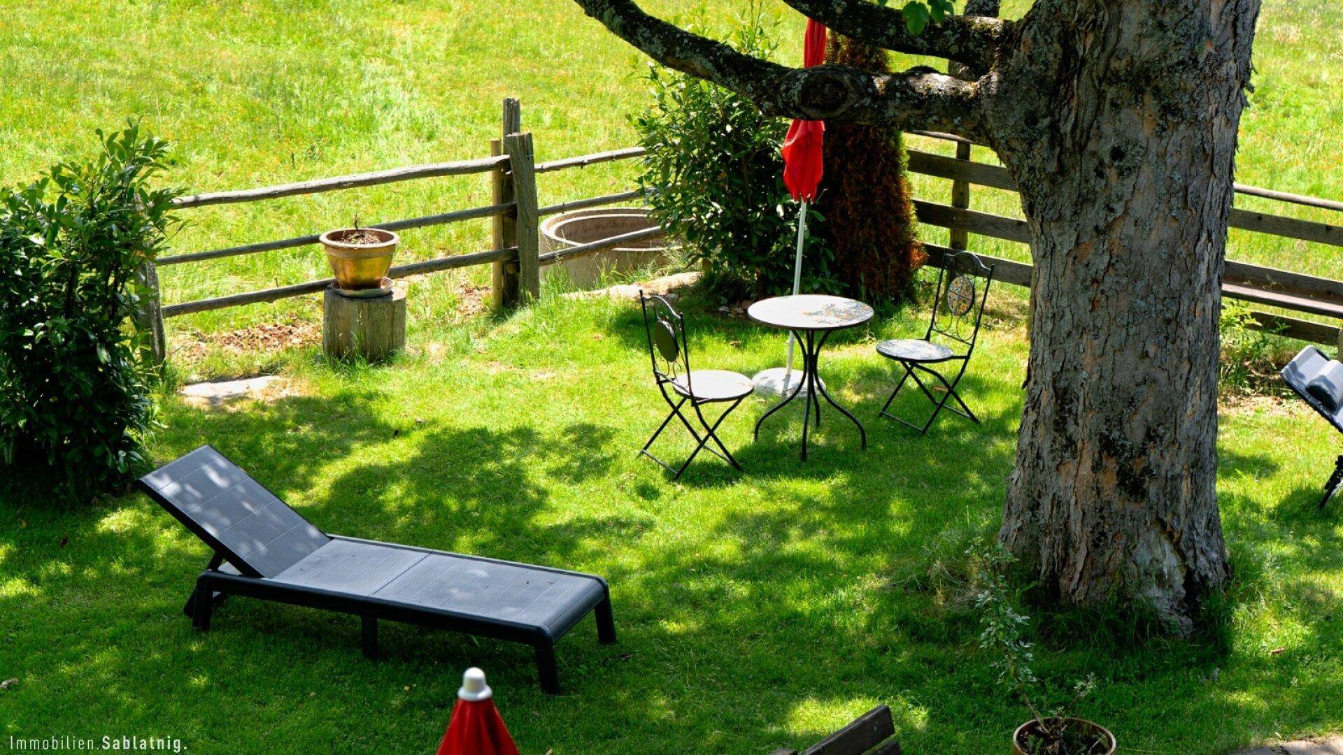 Entspannungsbereich im Garten mit Liegestuhl und kleinem Tisch unter einem schattenspendenden Baum.