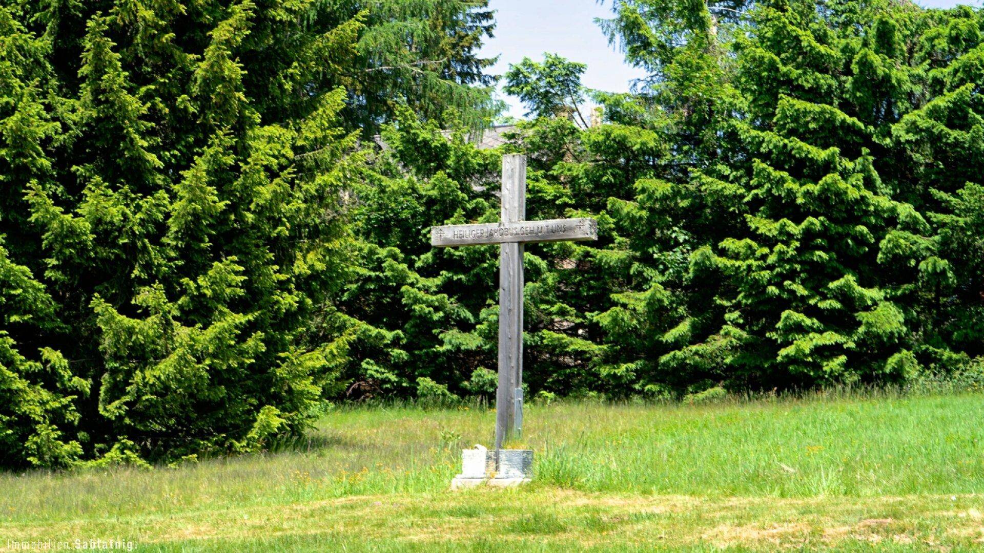 Ein schlichtes Holzkreuz steht auf einer grünen Wiese, umgeben von hohen Nadelbäumen.