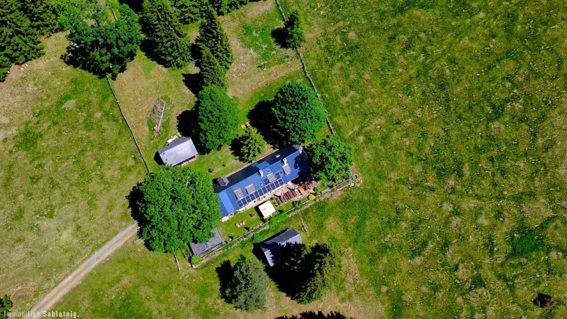 Luftaufnahme des Anwesens mit einem langen Haus, blauem Dach und Solarpaneelen, umgeben von Wiesen und Bäumen.