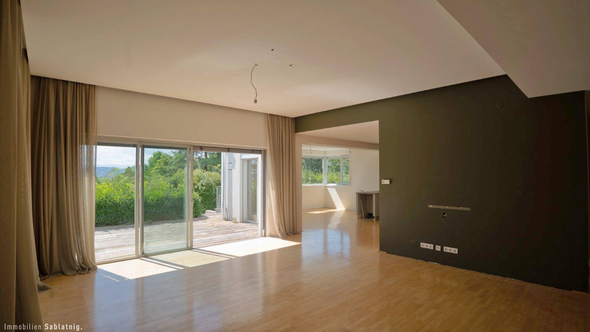 Geräumiges Wohnzimmer mit hellem Holzfußboden, großen Schiebetüren zur Terrasse und dunkler Akzentwand.