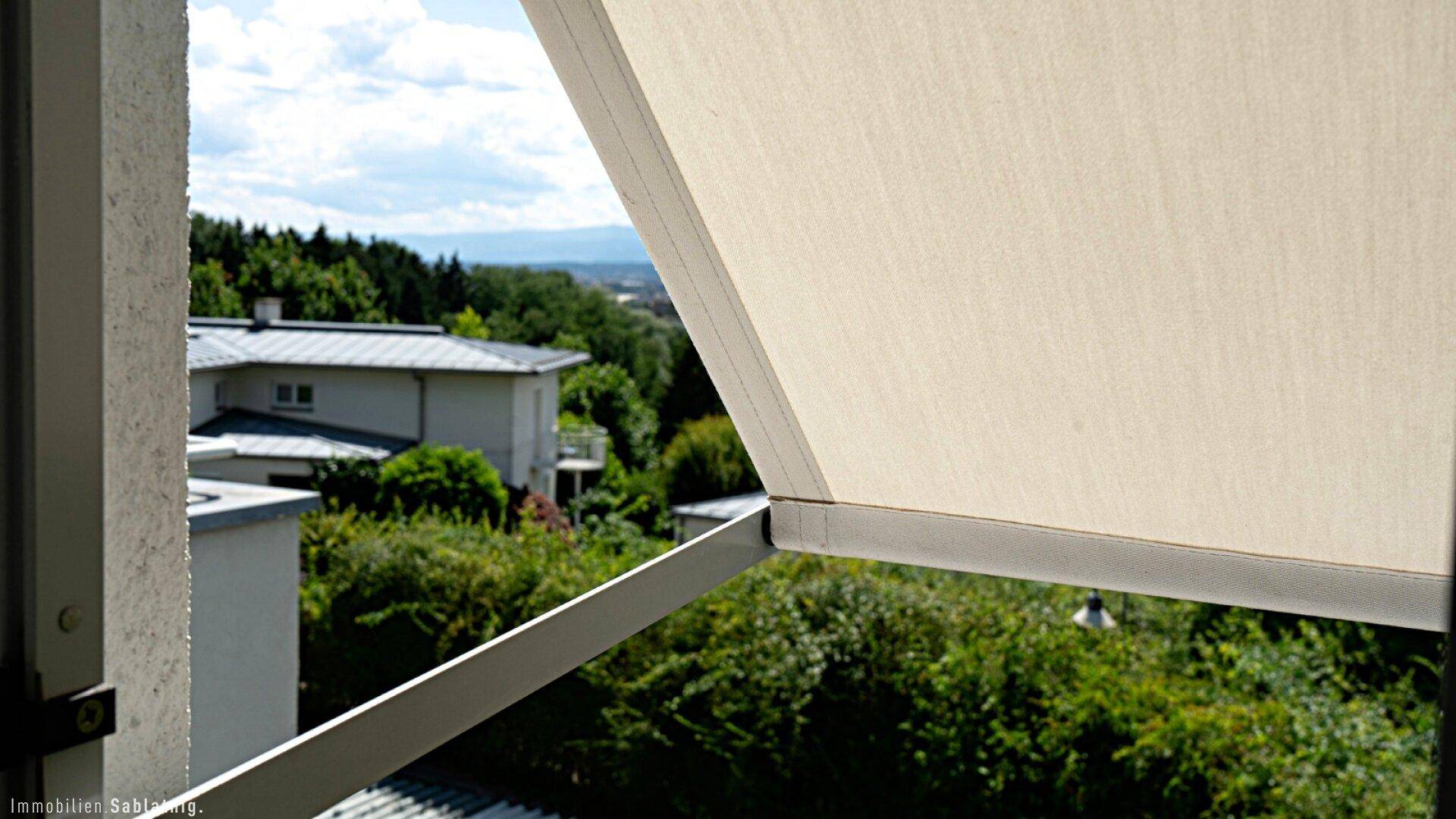 Ausblick aus einem Fenster mit Markise, zeigt entfernte Häuser und grüne Landschaft.