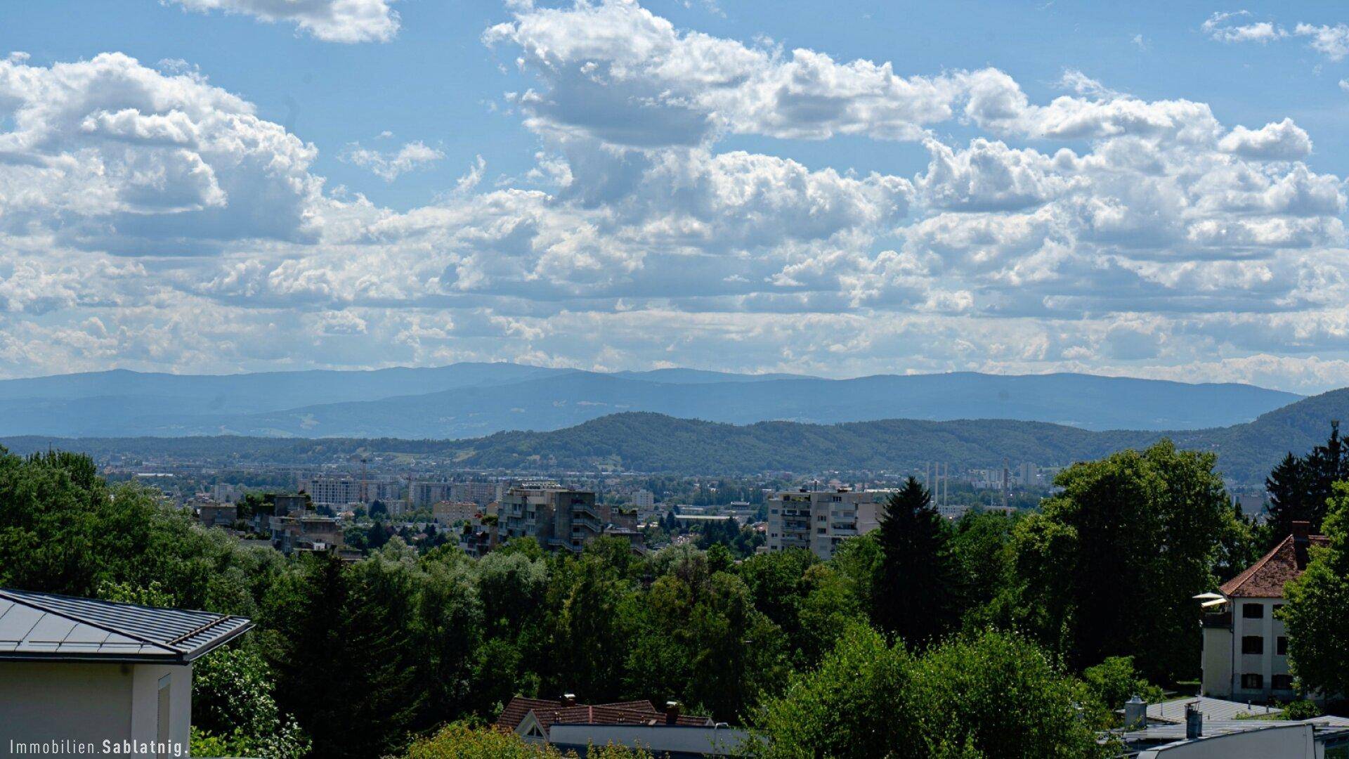 Panoramablick über eine Stadtlandschaft mit Bergen im Hintergrund unter blauem Himmel.