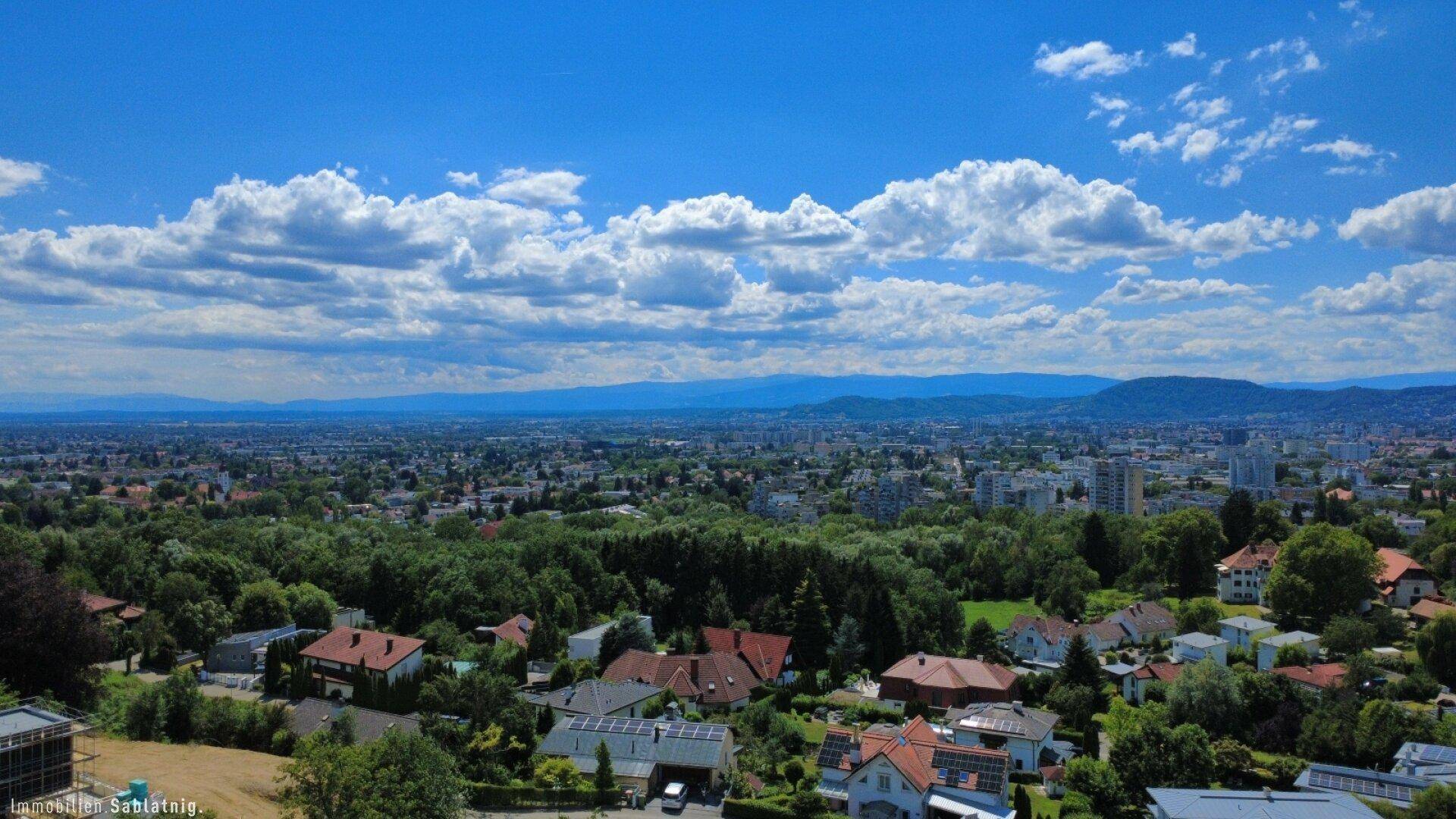 Panoramablick über eine weite Stadtlandschaft mit Bergen am Horizont unter einem blauen Himmel.