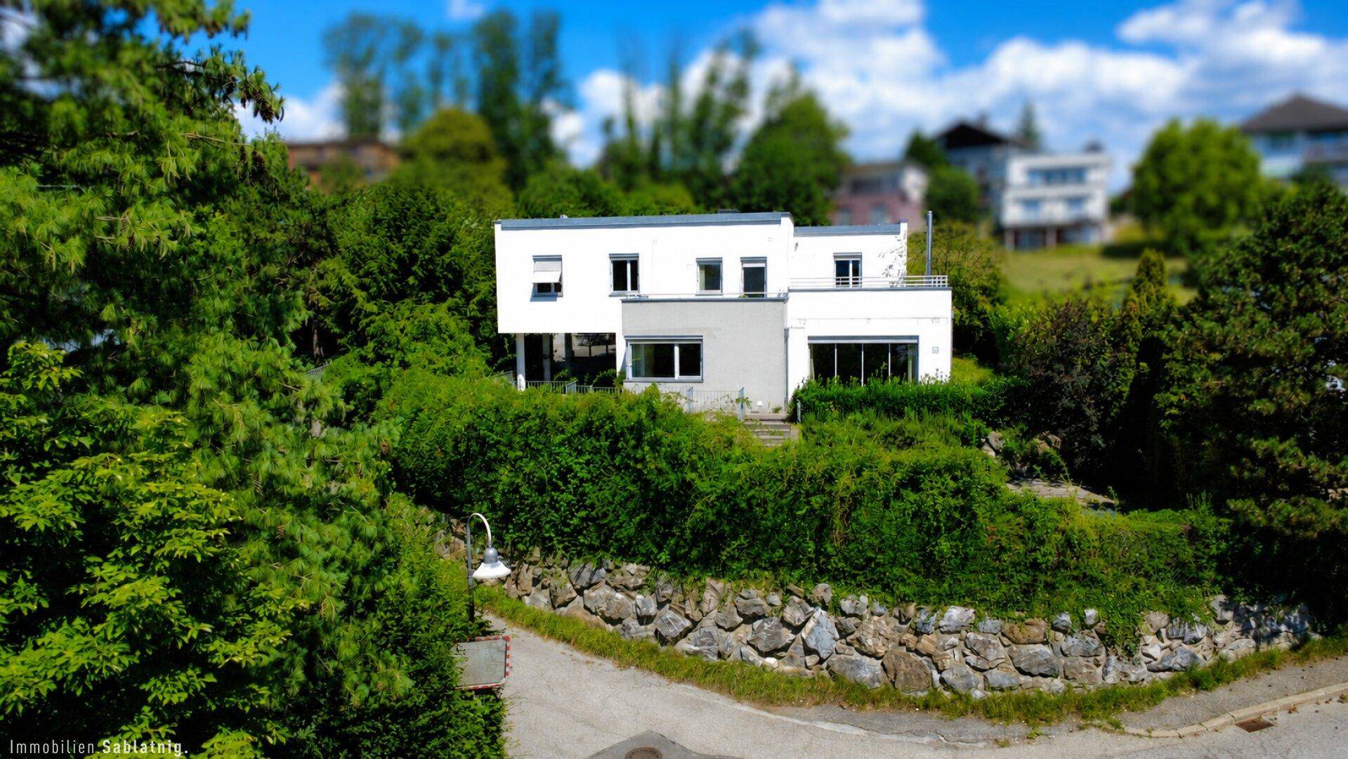 Modernes weißes Haus, umgeben von üppigem Grün und einer Natursteinmauer im Vordergrund.