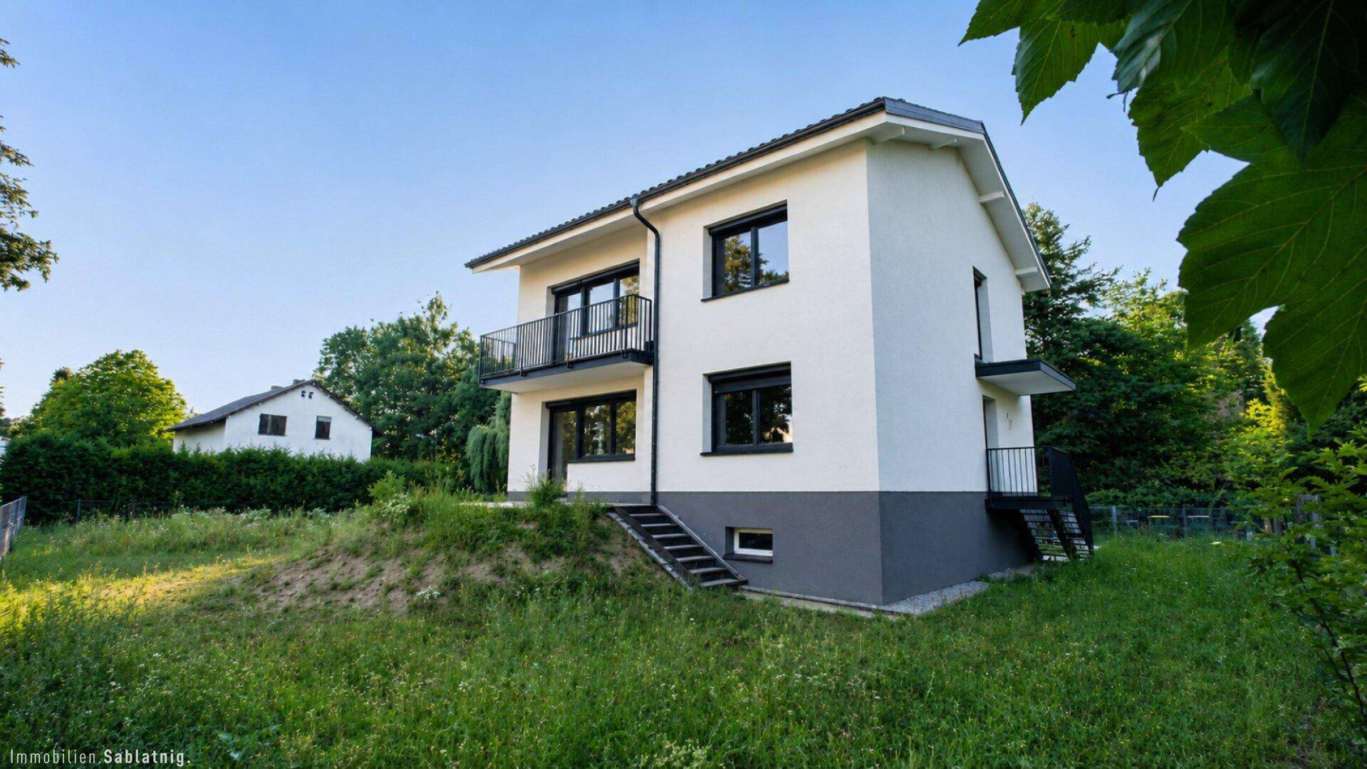 Freistehendes Einfamilienhaus mit weißer Fassade und dunklen Akzenten, umgeben von Grünflächen.