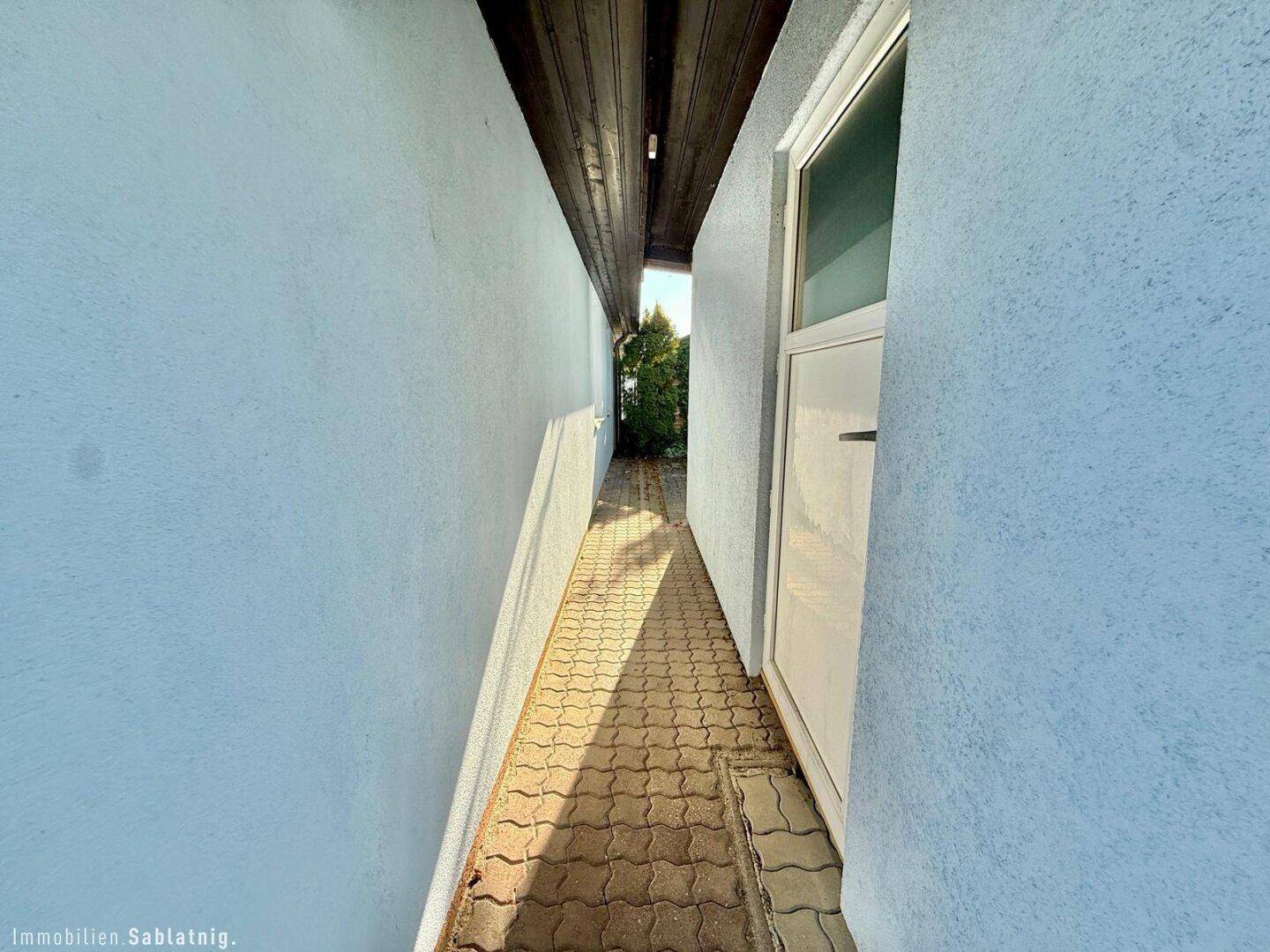 Schmaler gepflasterter Weg entlang der Hauswand, der zum Garten führt.