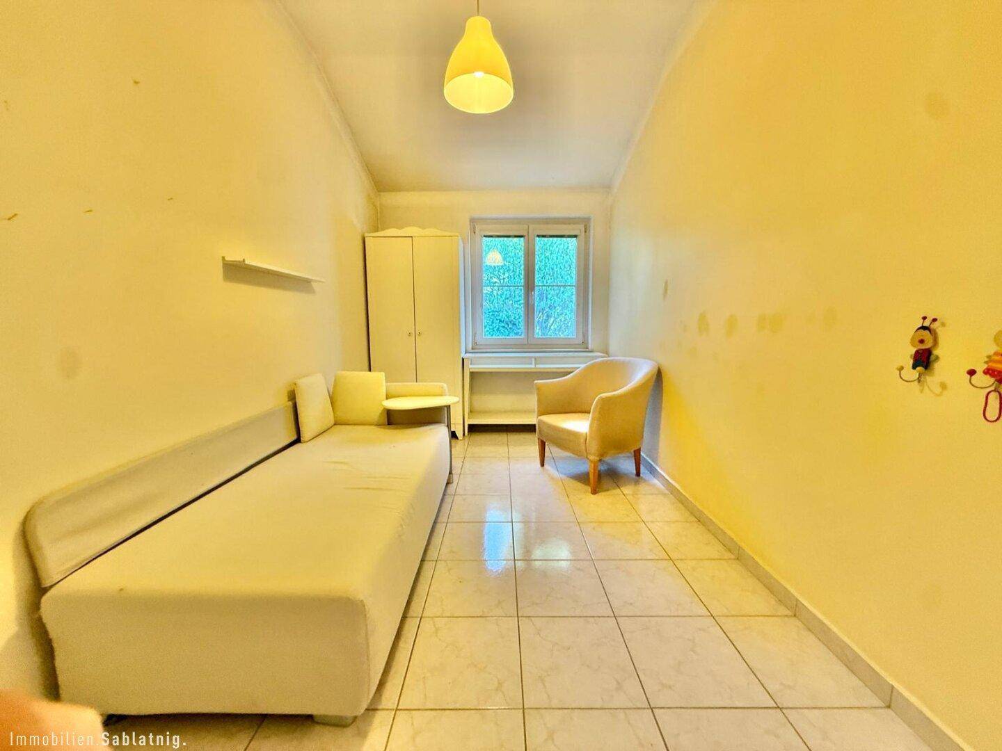 Helles Schlafzimmer mit Fliesenboden, Fenster und einfacher Einrichtung.