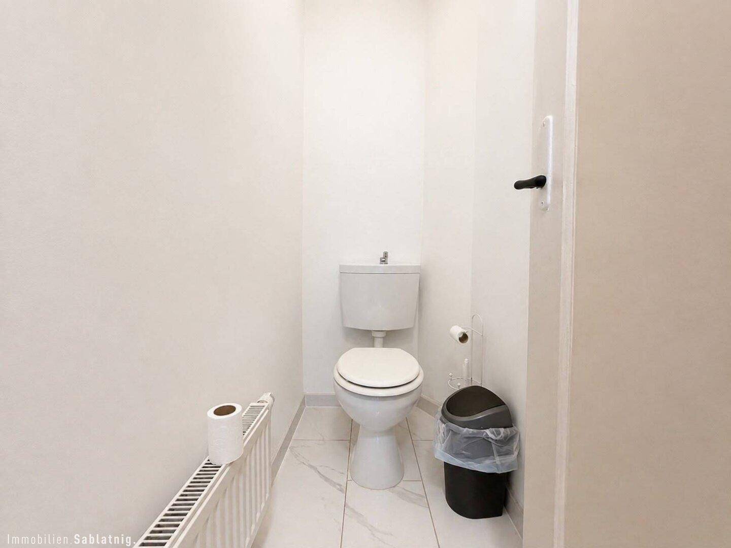 Separates WC mit weißer Toilette, Heizkörper und hellen Fliesen am Boden.