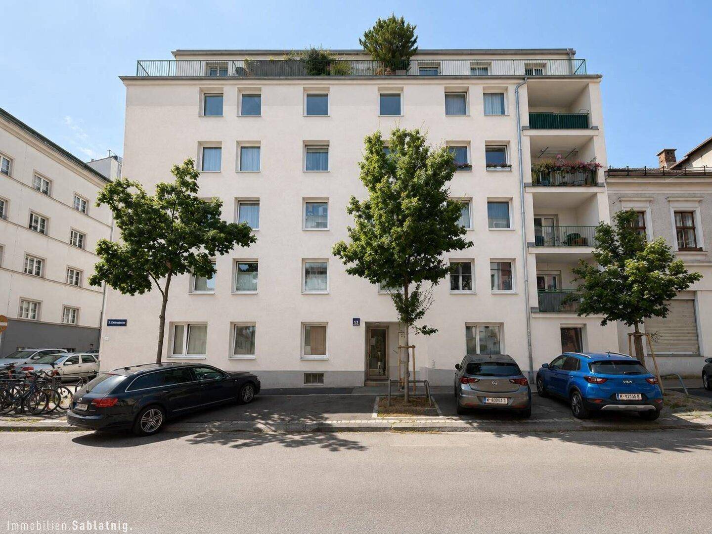 Mehrstöckiges Wohngebäude mit weißer Fassade, vielen Fenstern und Bäumen davor.