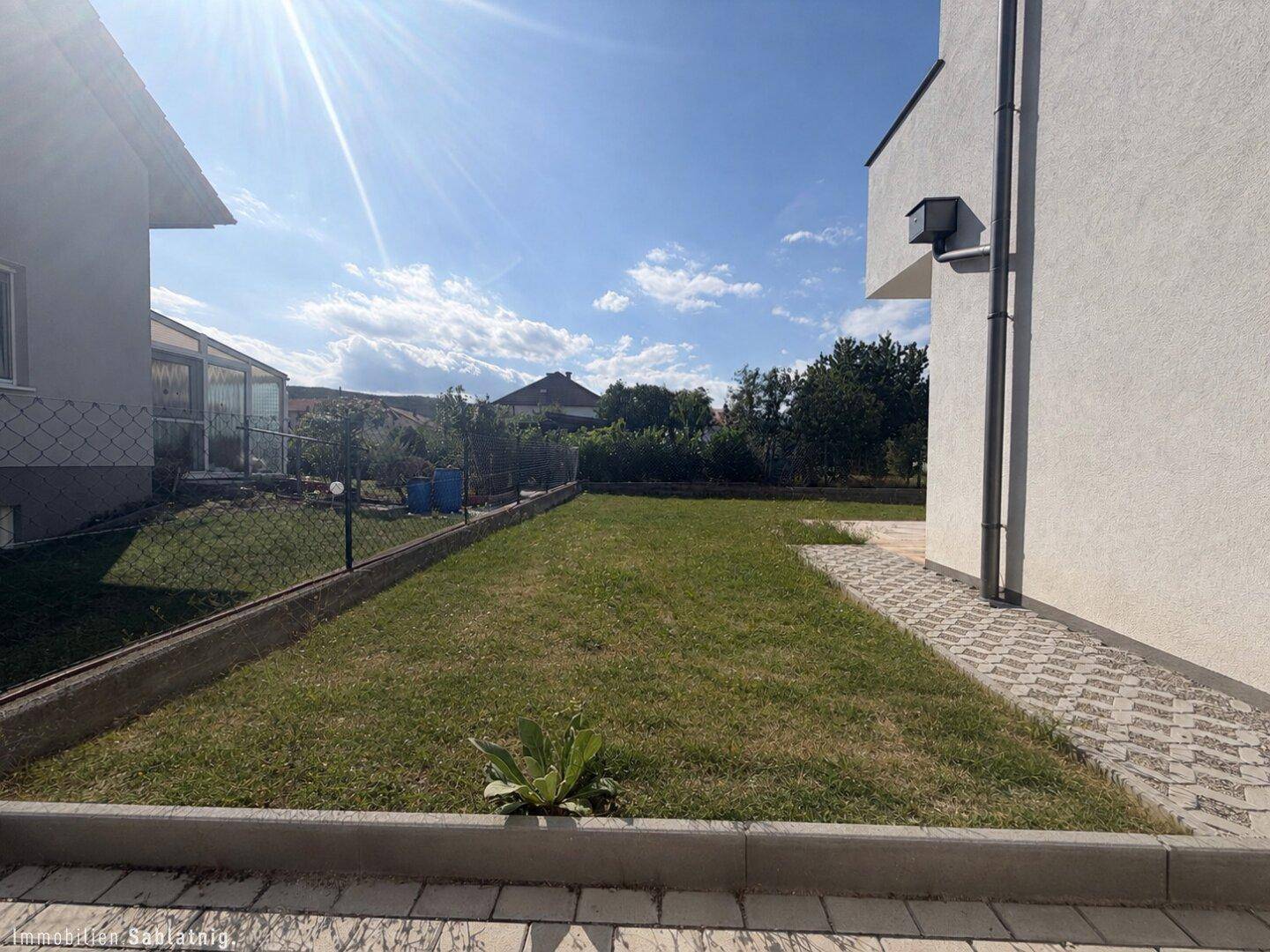 Sonniger Gartenbereich mit Rasenfläche und gepflastertem Weg neben dem modernen Haus.