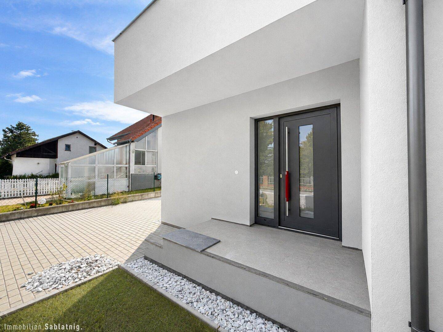 Moderner Hauseingang mit dunkler Tür, gepflastertem Vorplatz und Kiesbeet.