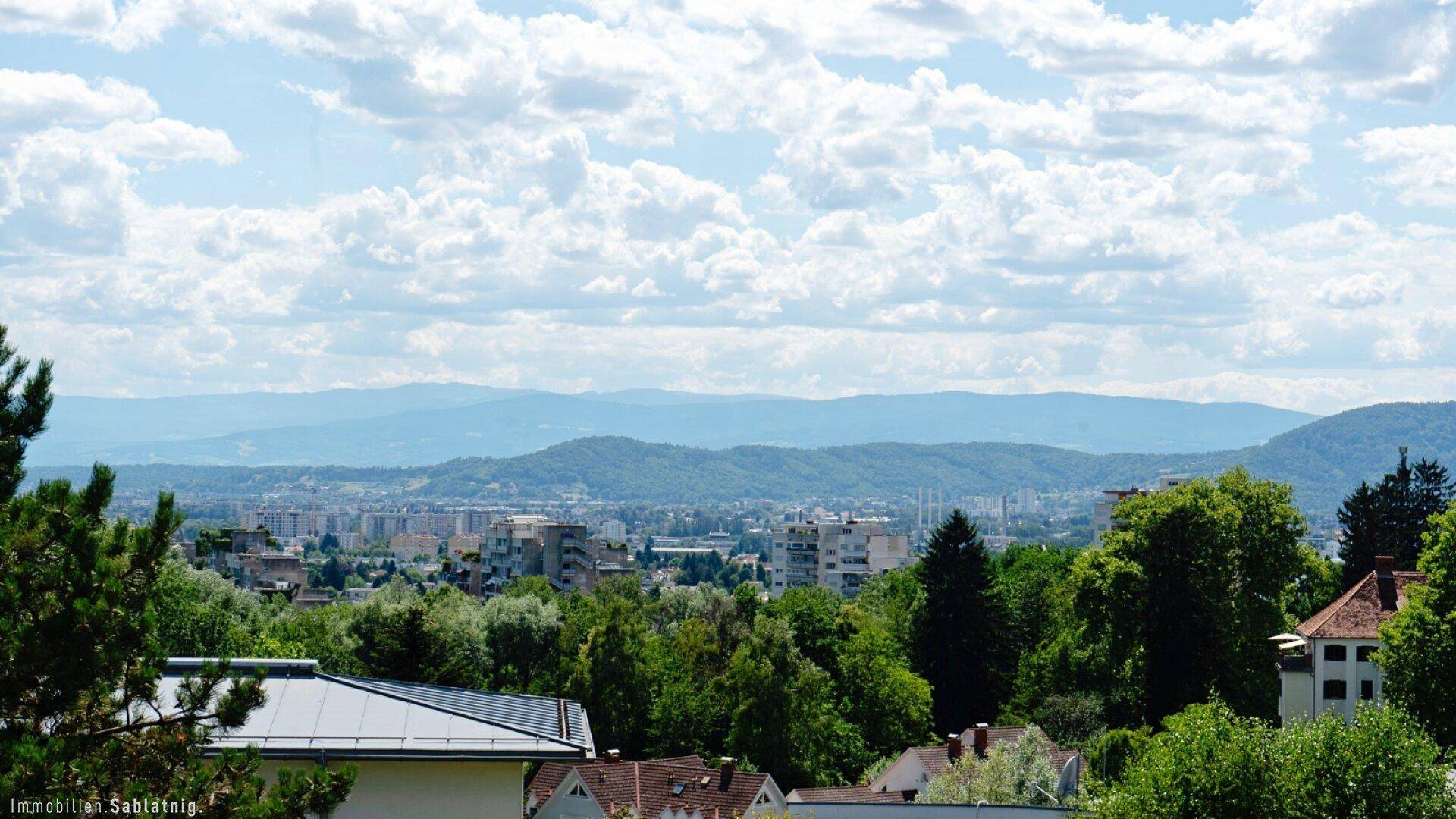 Weitläufiger Blick über die Stadt und die umliegende Berglandschaft unter einem bewölkten Himmel.