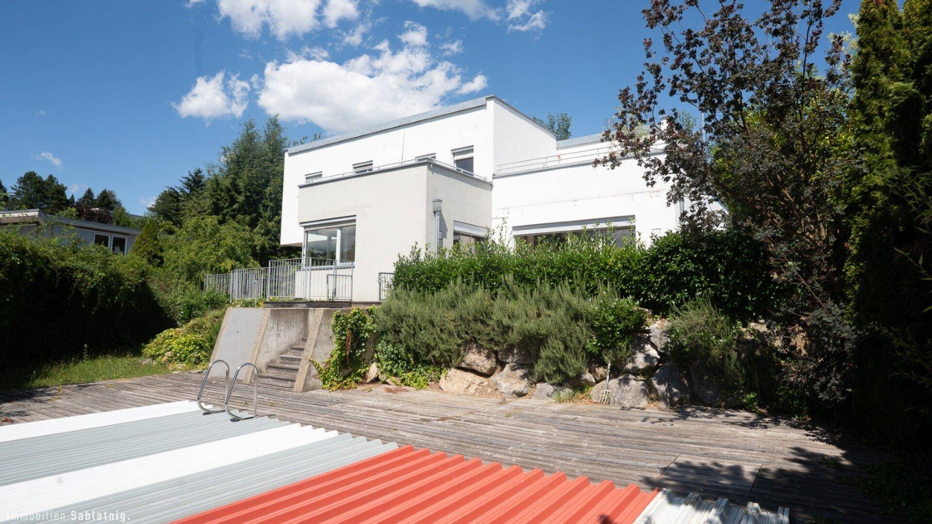 Modernes Haus mit abgedecktem Swimmingpool und Holzdeck, umgeben von üppigem Grün.