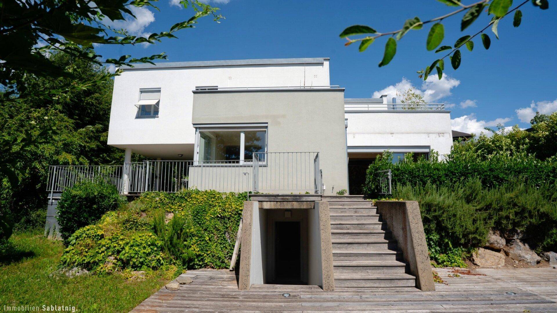 Moderne Hausfassade mit Treppe zum Eingangsbereich, umgeben von üppiger Vegetation.