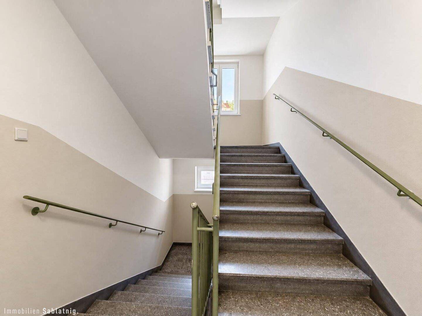 Helles Treppenhaus mit Steinstufen, grünen Handläufen und Fenstern.