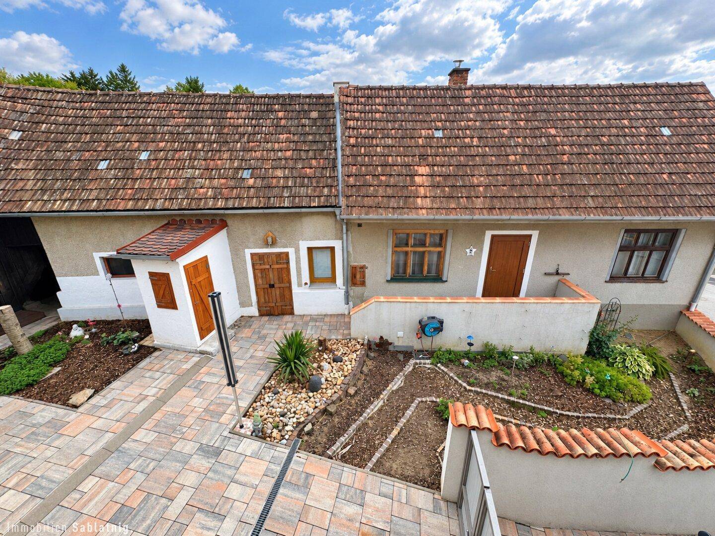 Außenbereich mit gepflastertem Weg, Gartenbeeten und traditionellen Gebäuden im Hintergrund.