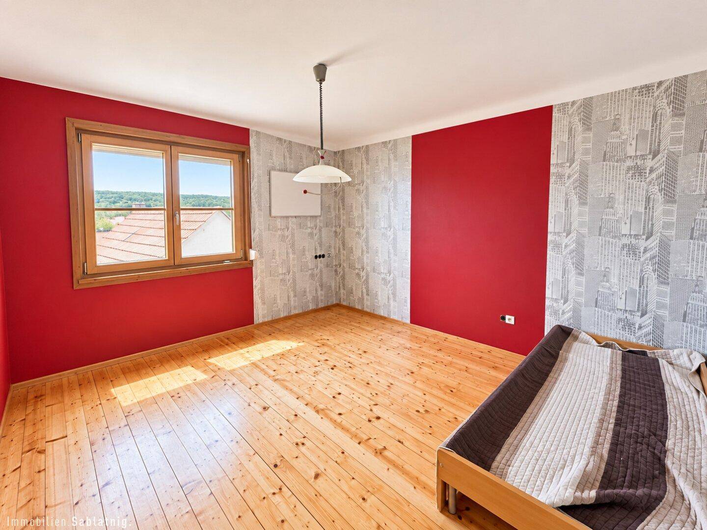 Helles Schlafzimmer mit Holzboden, roten Wänden und einem Fenster mit Blick ins Grüne.