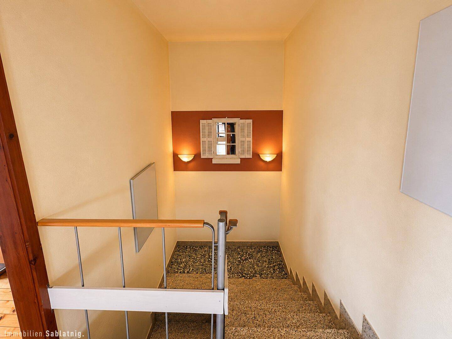 Treppenhaus mit Terrazzoboden, Holzgeländer und dekorativer Wandbeleuchtung.
