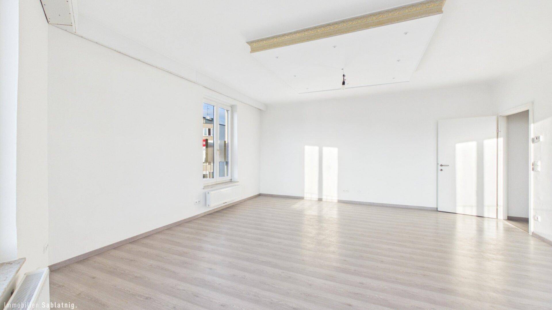 Helles, leeres Zimmer mit Laminatboden und einem Fenster, das viel Tageslicht hereinlässt.