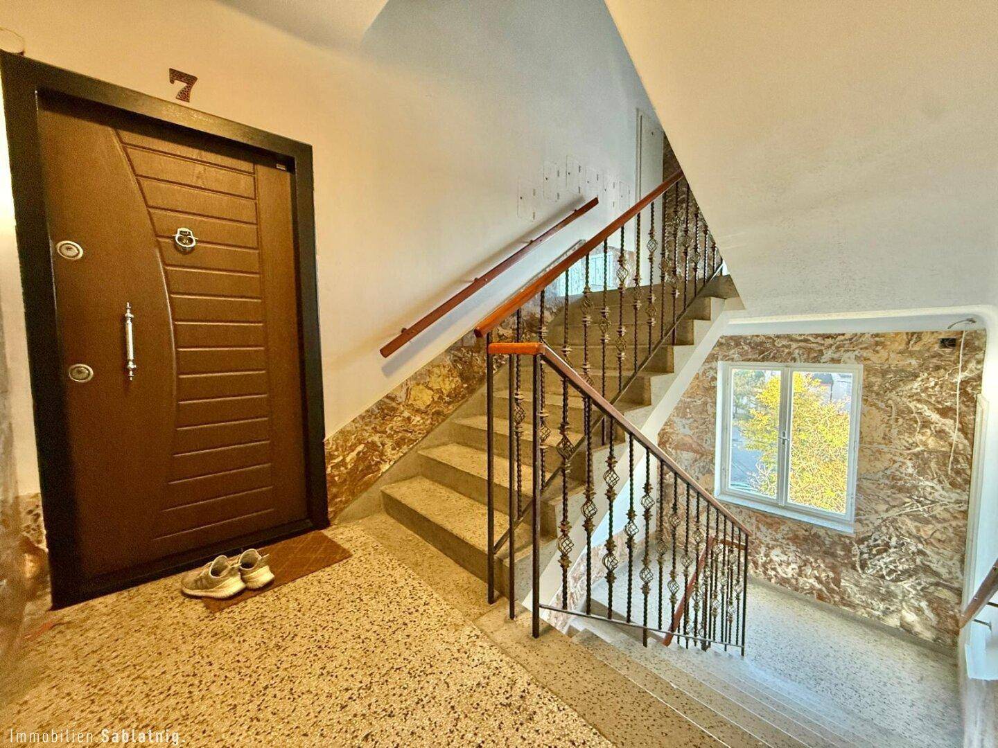 Treppenhaus mit dunkler Eingangstür, schmiedeeisernem Geländer und Terrazzoboden.
