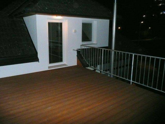 Holzterrasse im Obergeschoss mit Metallgeländer und Zugang zum Innenbereich bei Nacht.