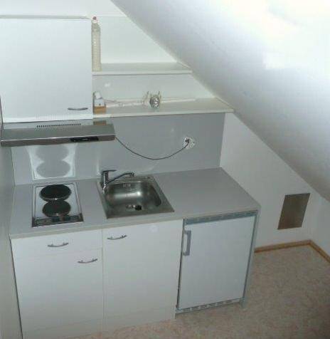 Kleine Küchenzeile im Dachgeschoss mit Kochfeld, Spüle, Kühlschrank und weißen Schränken.