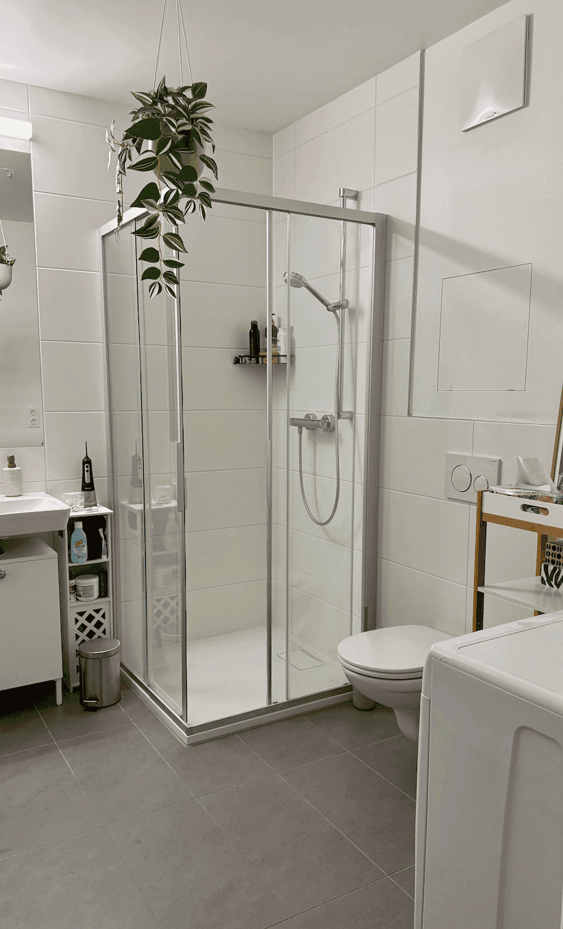 Modernes Badezimmer mit Duschkabine, WC, Waschbecken und Waschmaschine auf grauen Fliesen.