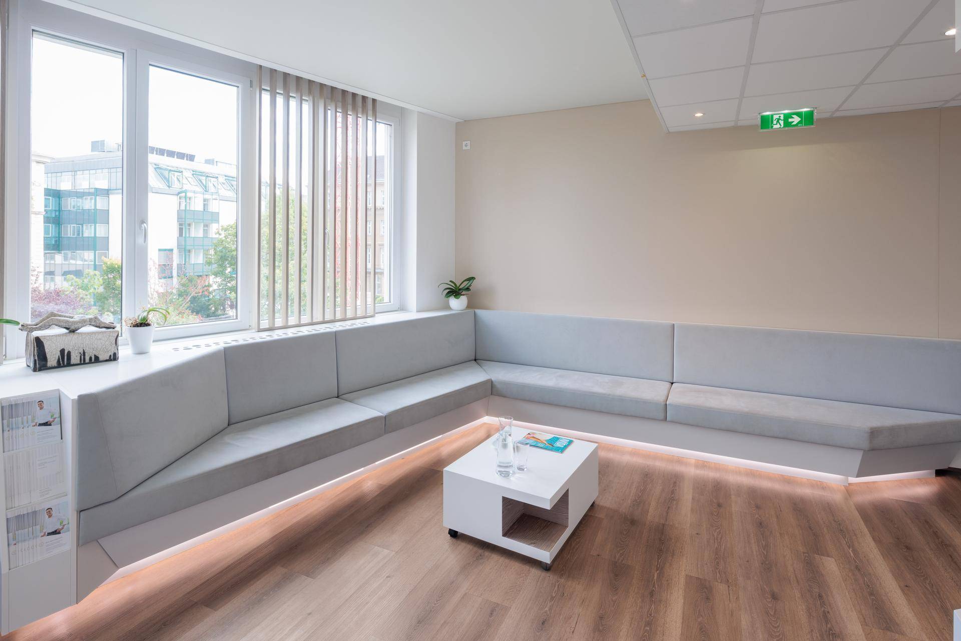 Heller Wartebereich mit bequemer L-förmiger Sitzbank, kleinem Couchtisch und großen Fenstern mit Jalousien.