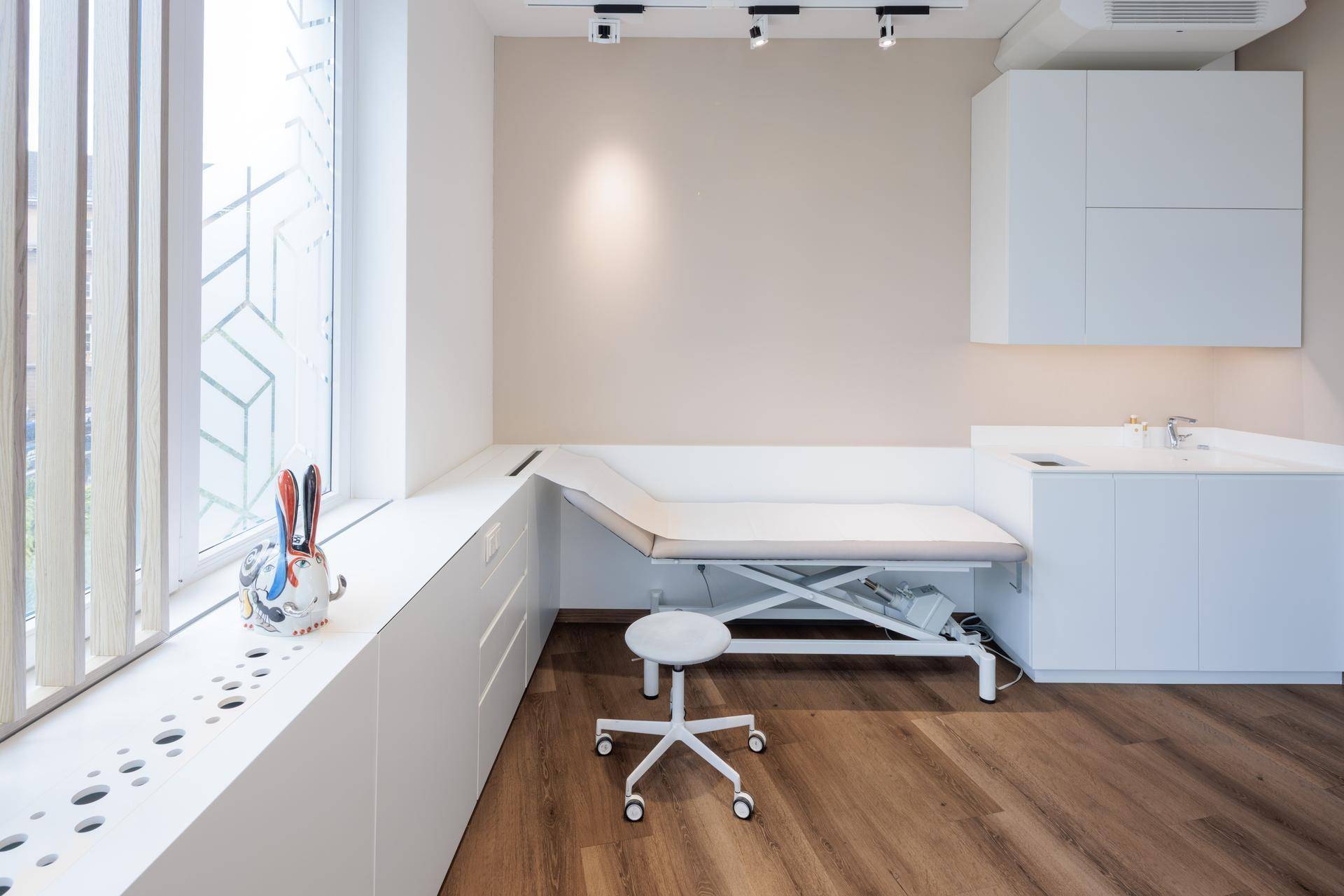 Modernes Behandlungszimmer mit Untersuchungsliege, Waschbecken und weißen Einbauschränken unter einem Fenster.