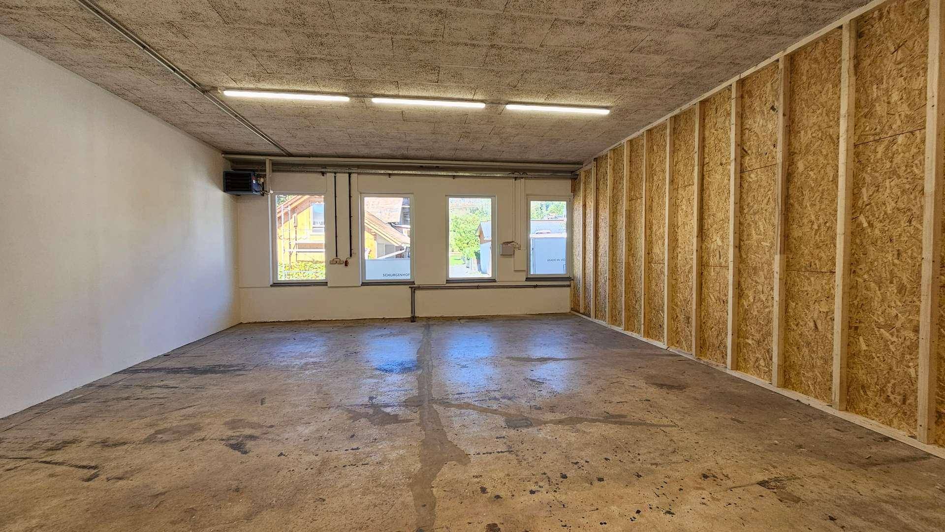 Großer, leerer Gewerberaum mit Betonboden, sichtbarer Decke, Holzverkleidung und Fenstern für Tageslicht.
