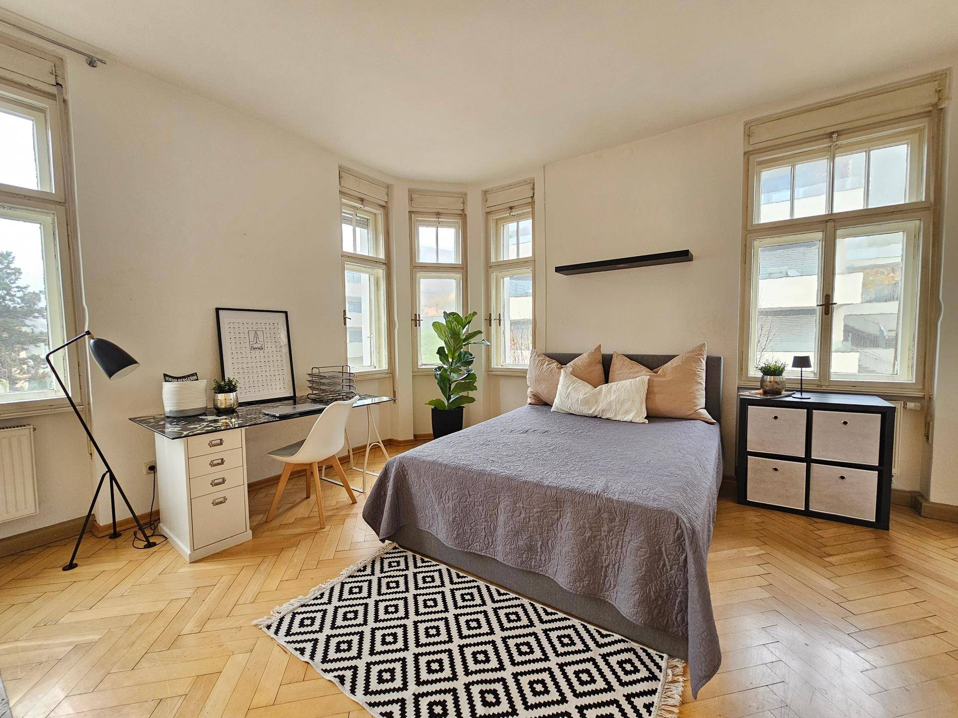 Geräumiges Schlafzimmer mit vielen Fenstern, Fischgrätparkett und einem Arbeitsbereich.