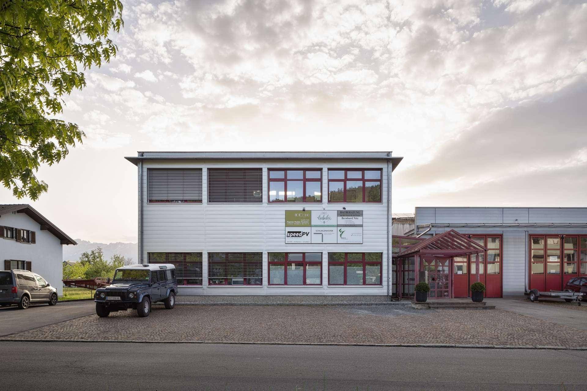 Ansicht des modernen Geschäftsgebäudes mit roten Fensterrahmen und Parkplätzen unter bewölktem Himmel.